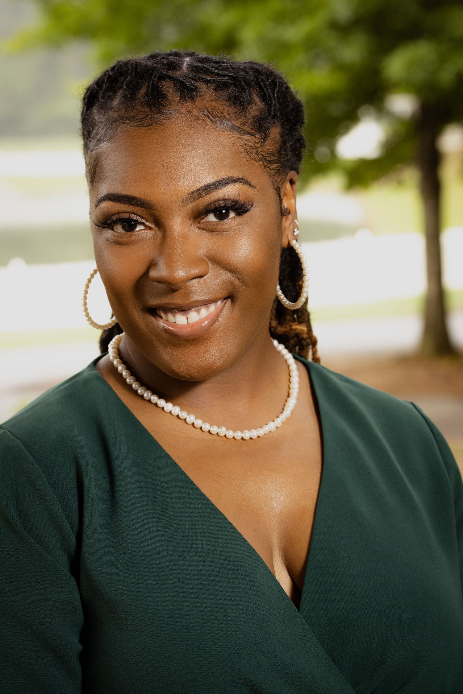 Smiling young woman wearing a green wrap top, pearl necklace, and gold hoop earrings, posing outdoors near a pond.