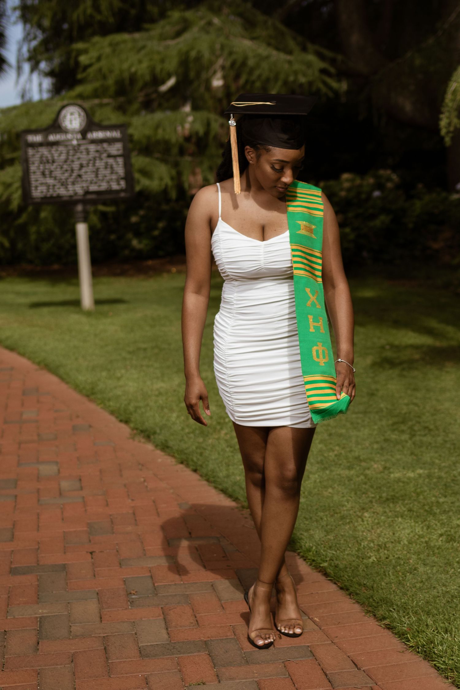 Young woman in white dress and graduation cap walks brick path wearing green Greek letter sash outdoors.