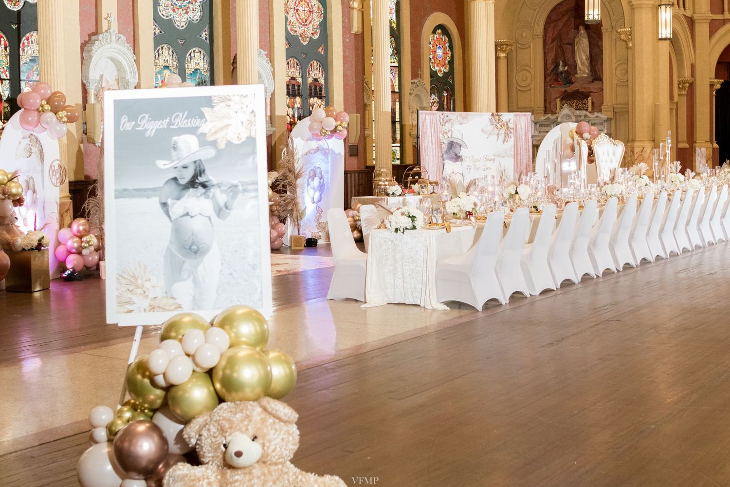 Elegant wedding reception ballroom with white chairs and long tables decorated in gold and blush pink accents.