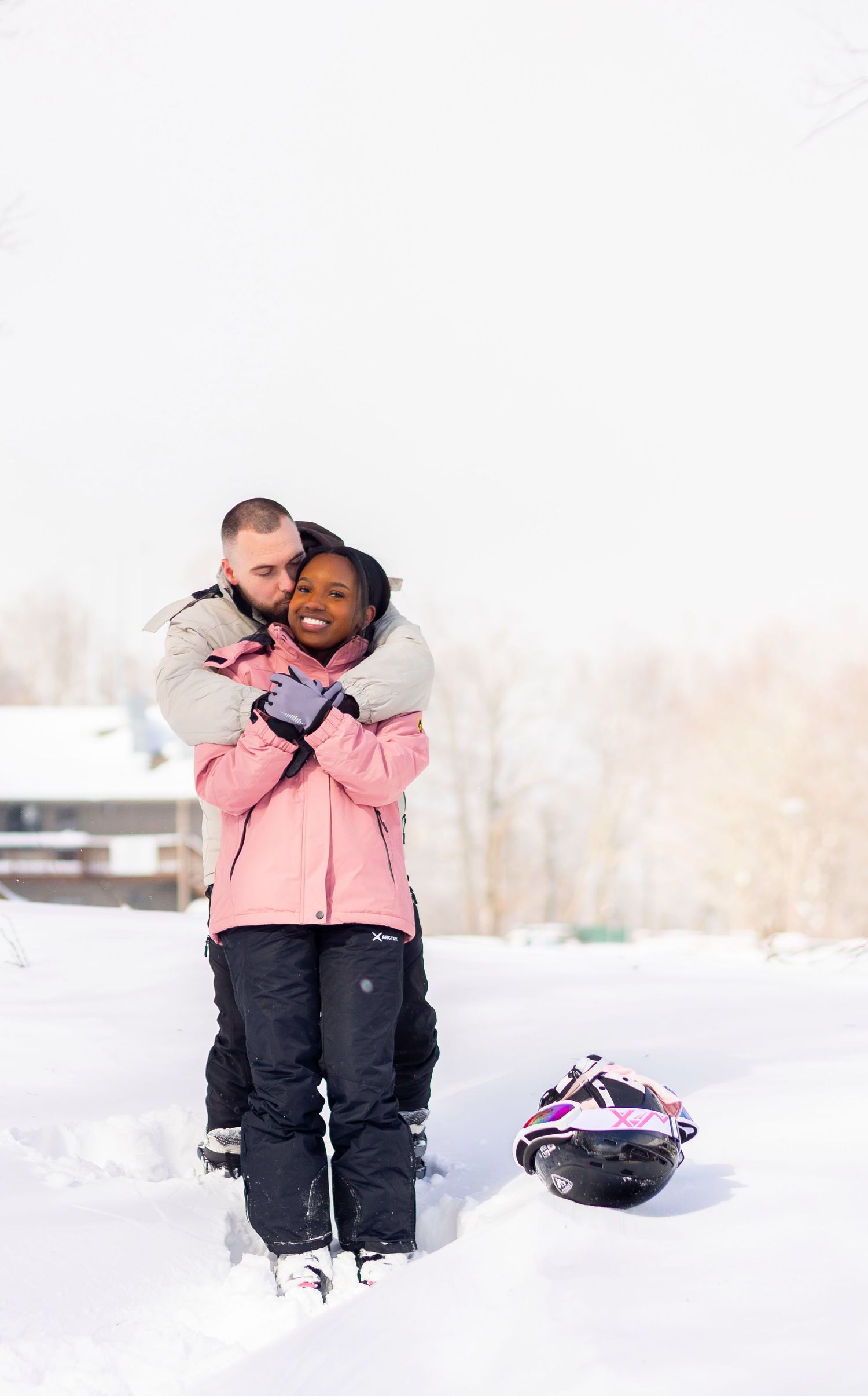 Couple embracing in snow wearing ski gear, woman in pink jacket smiling, helmet on ground nearby.