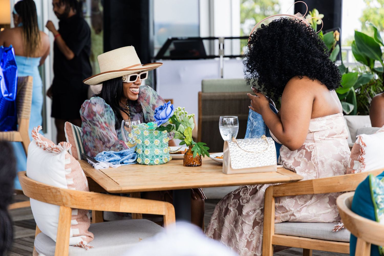 Two people enjoy an afternoon at an outdoor cafe table with drinks and flowers on a sunny day.