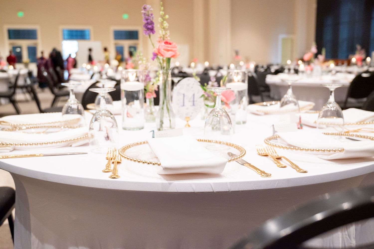 Elegant wedding reception table with white linen, gold chargers, crystal glasses, and floral centerpieces in a banquet hall.