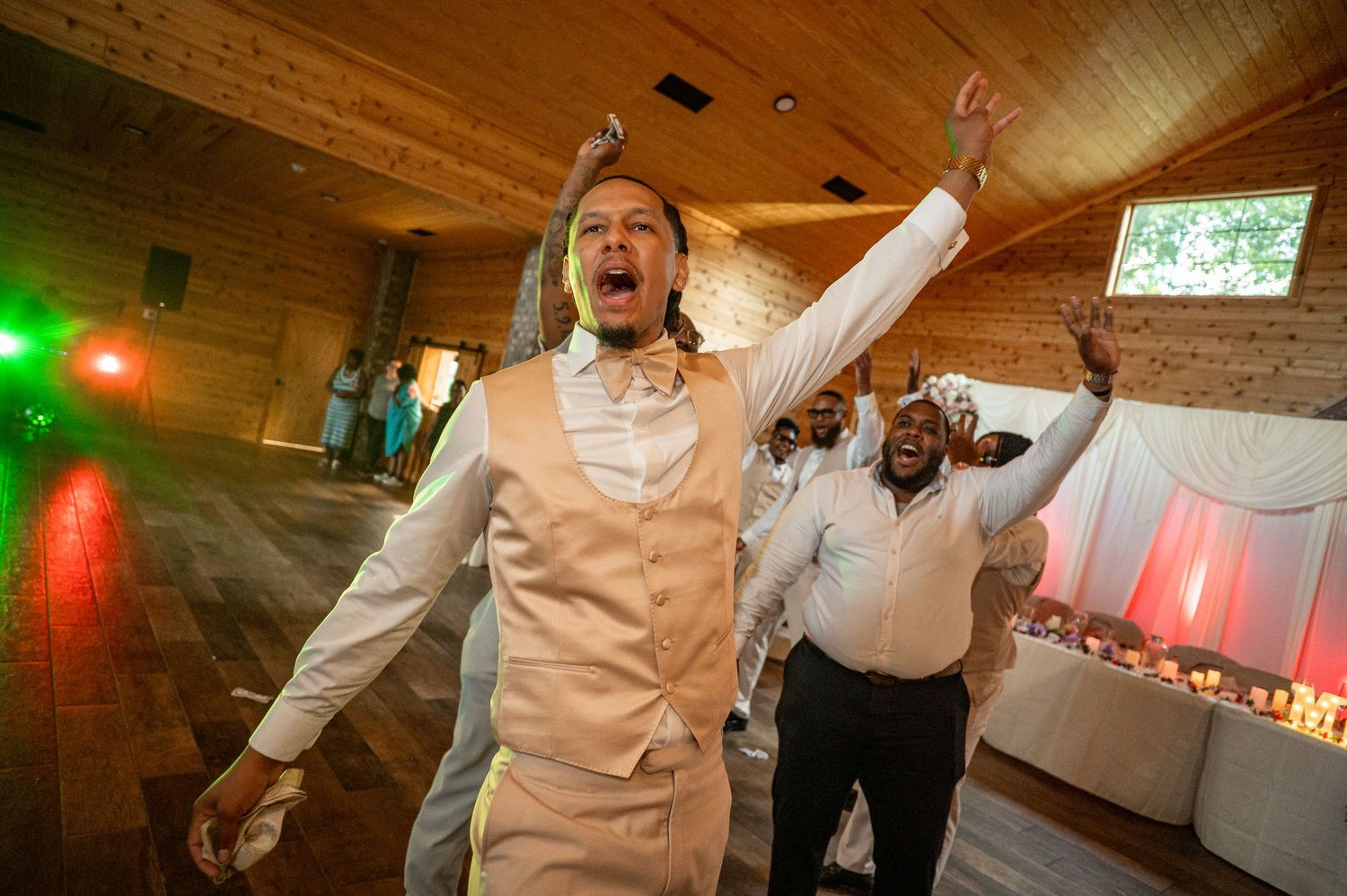 Excited groom in beige vest celebrates with raised arms on wedding reception dance floor with guests.
