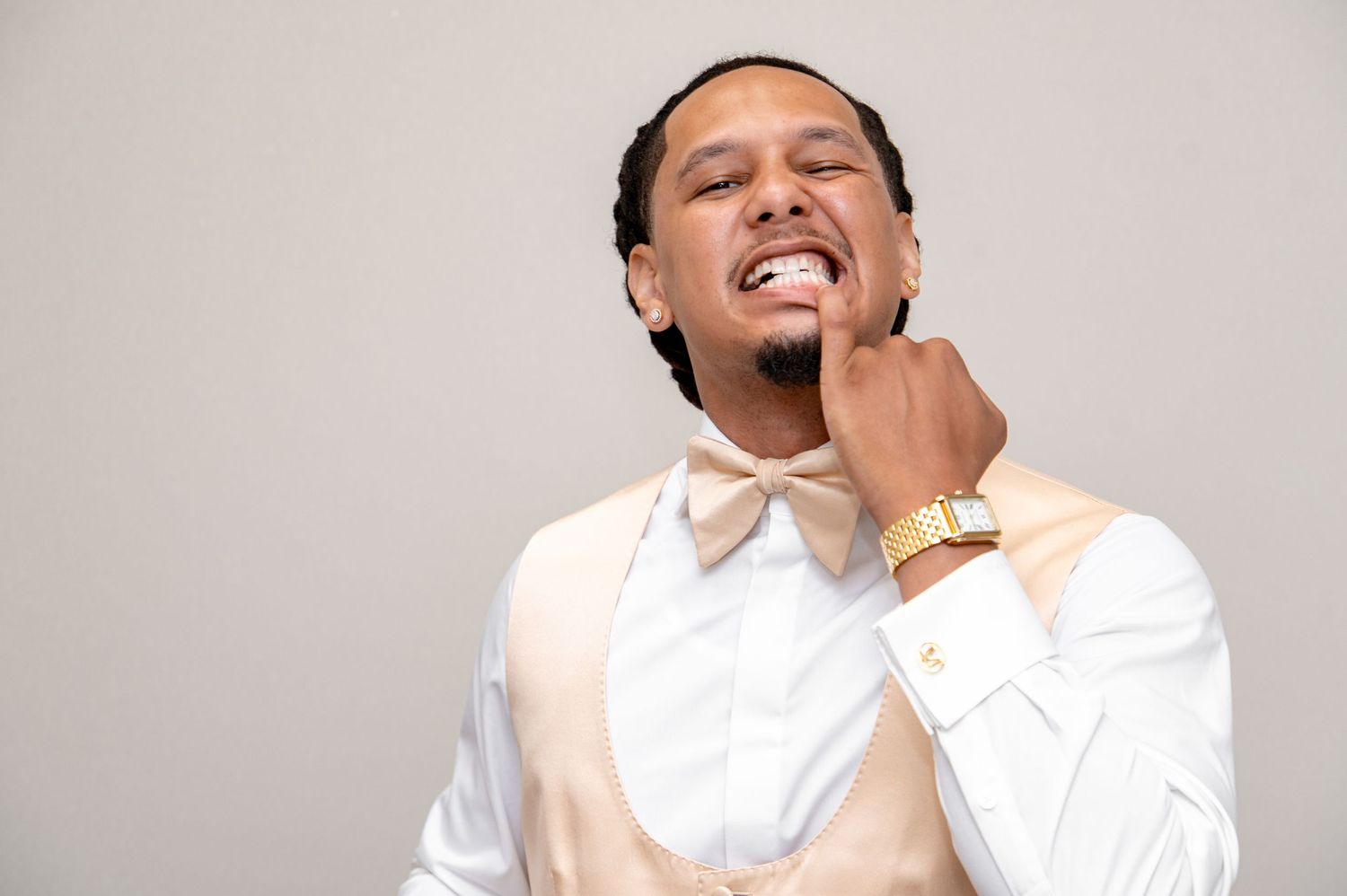 Smiling groom in cream vest and white shirt with gold bow tie and watch posing for portrait.