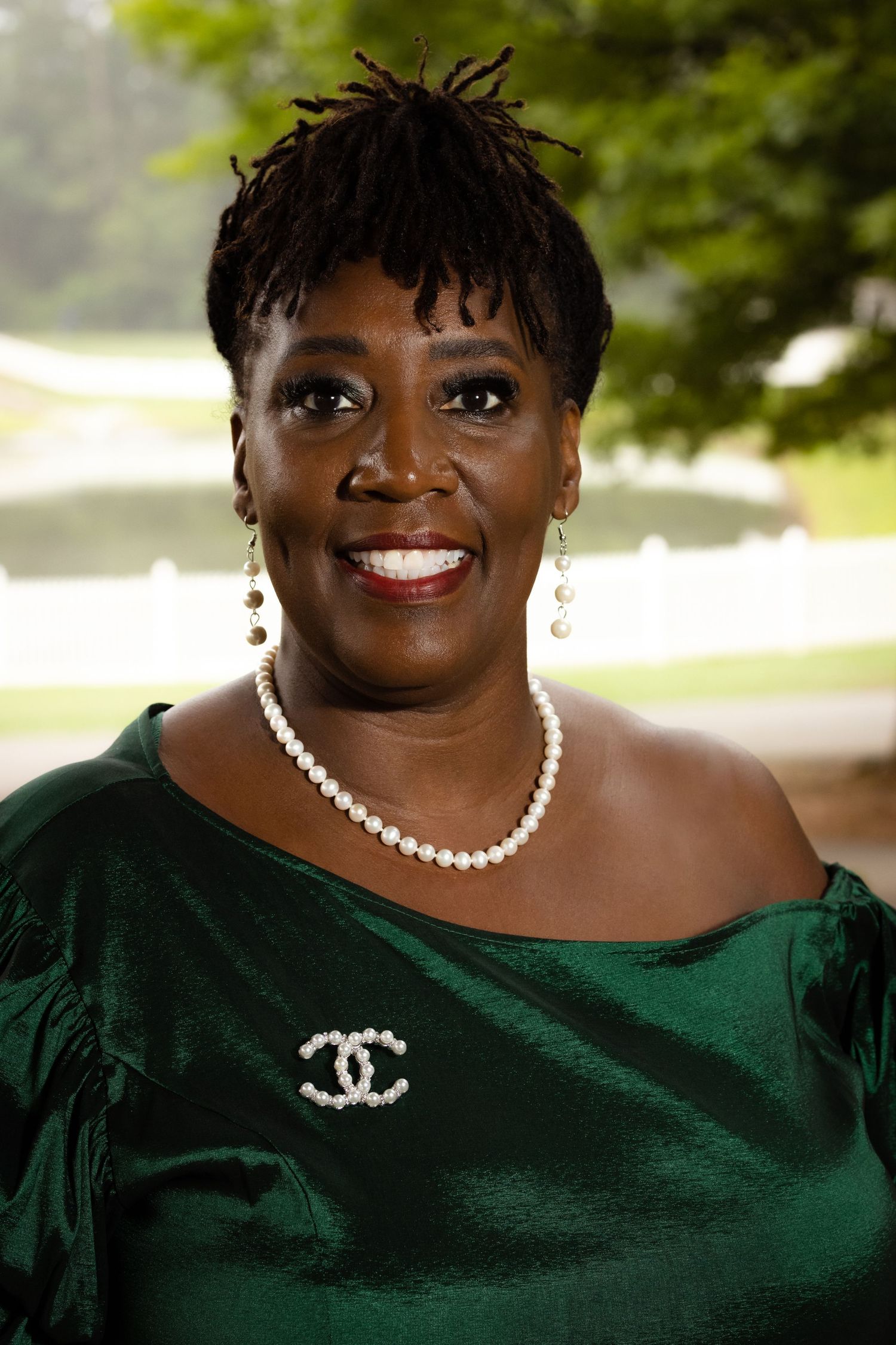 Smiling woman wearing a green satin off-shoulder dress, pearl necklace, and Chanel brooch outdoors.