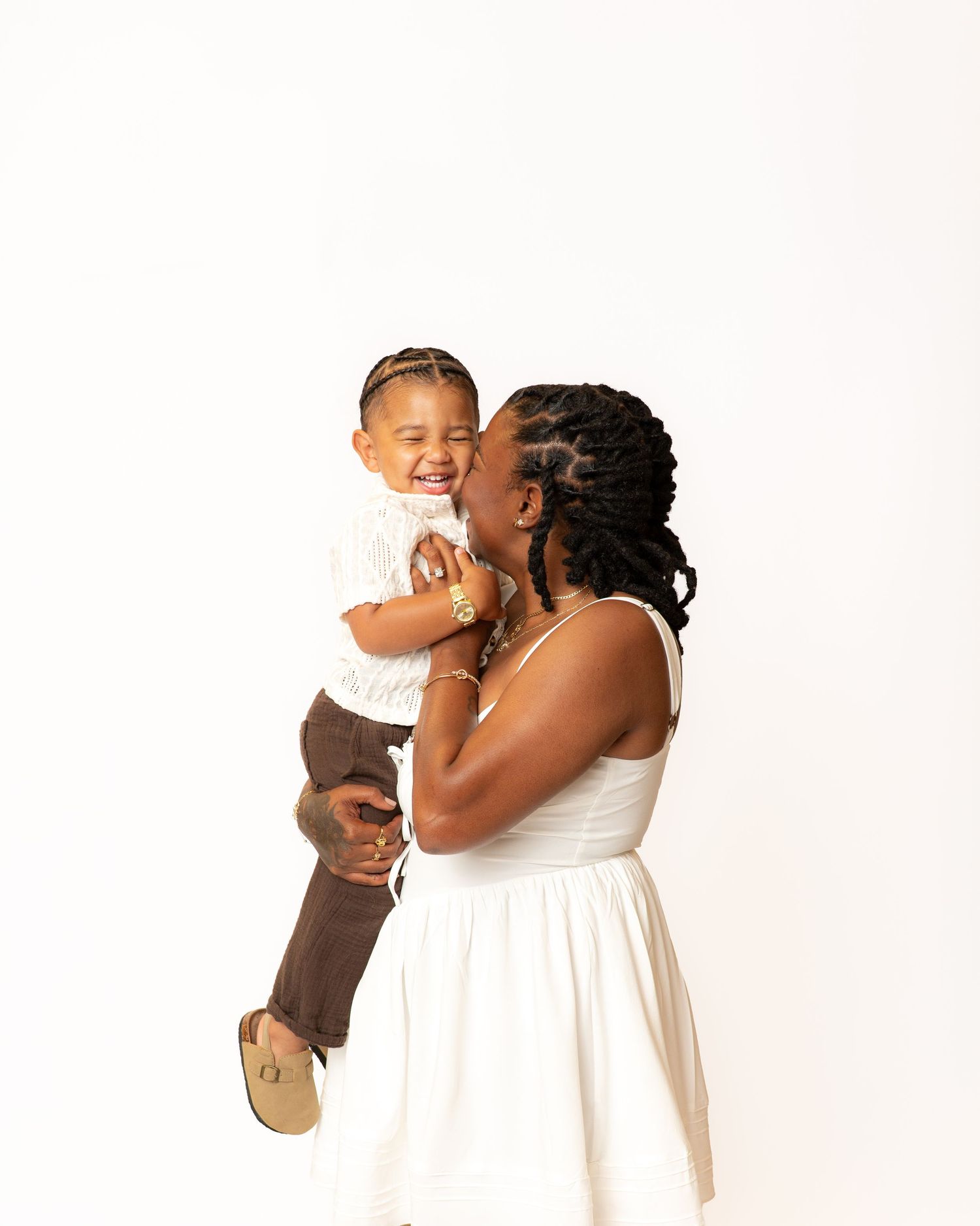 A joyful mother in a white dress kisses her laughing toddler boy in a bright studio portrait.