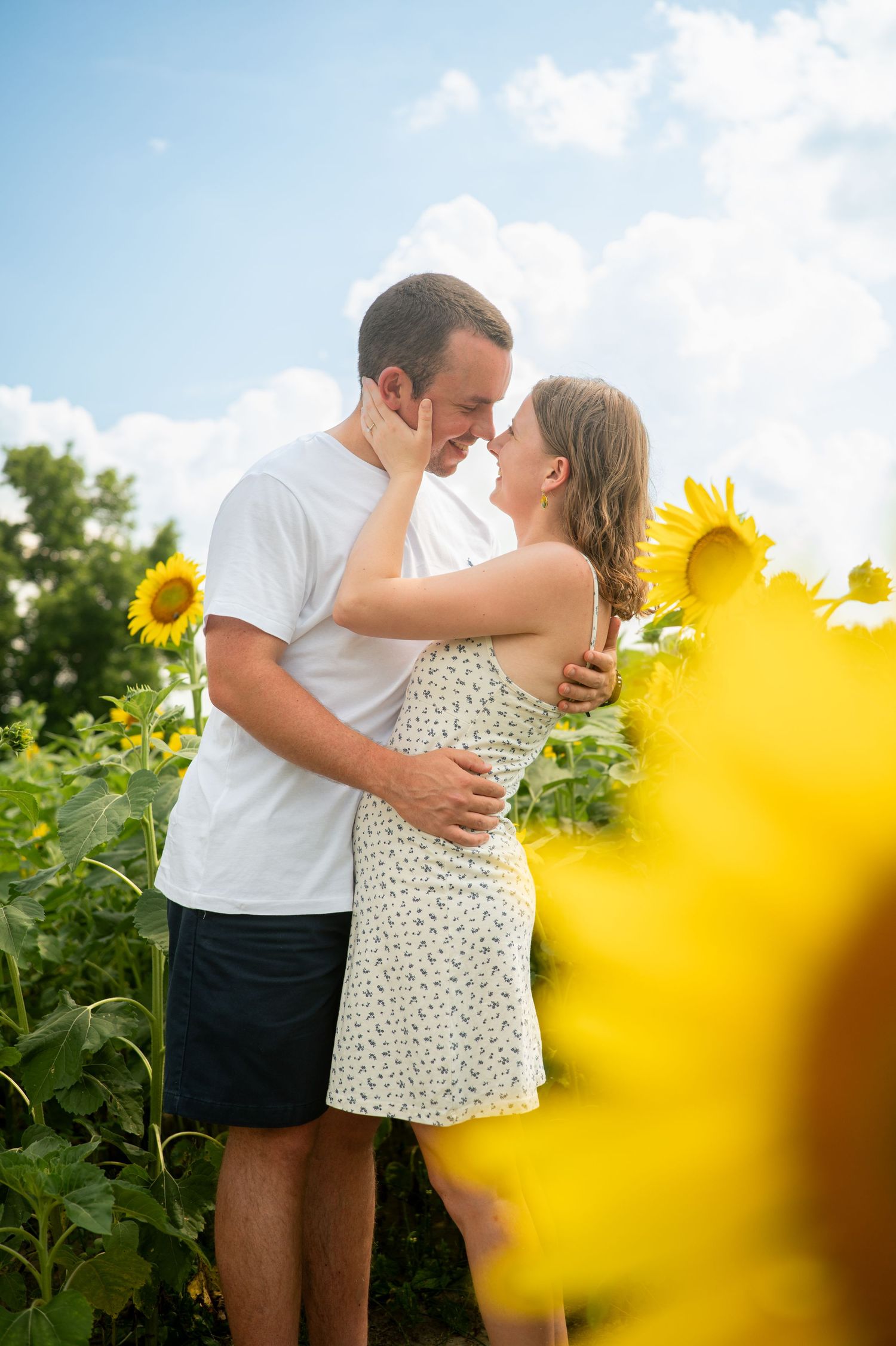 Couple sharing a romantic moment in a sunny sunflower field, embracing and about to kiss under a blue sky.