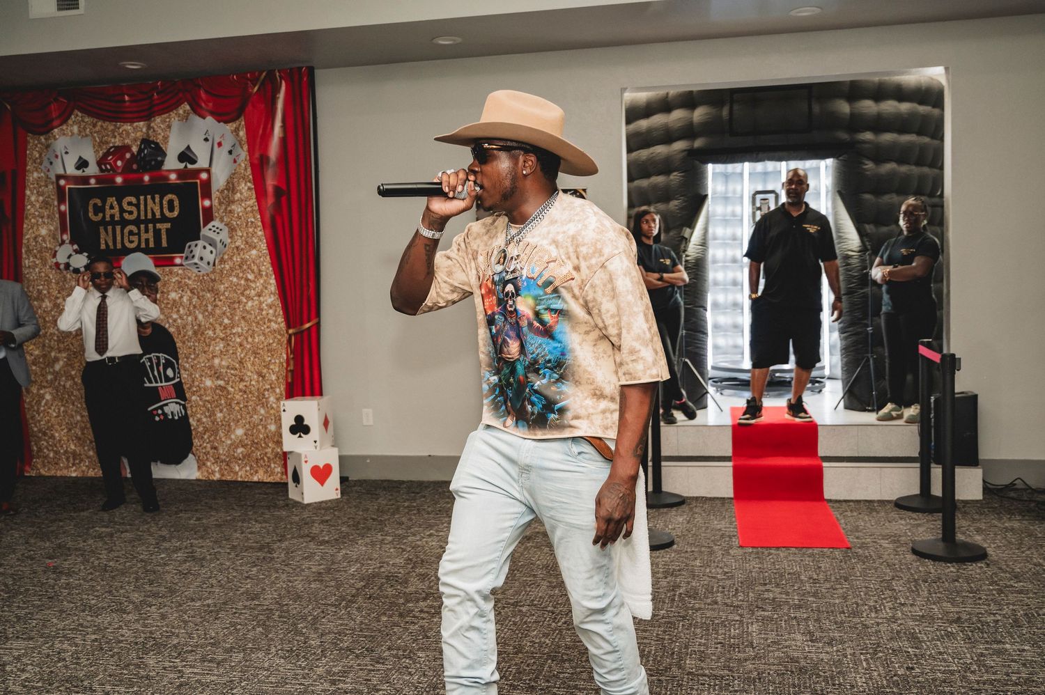 Performer in casual western attire entertains crowd at indoor venue with red carpet and decorative backdrop.