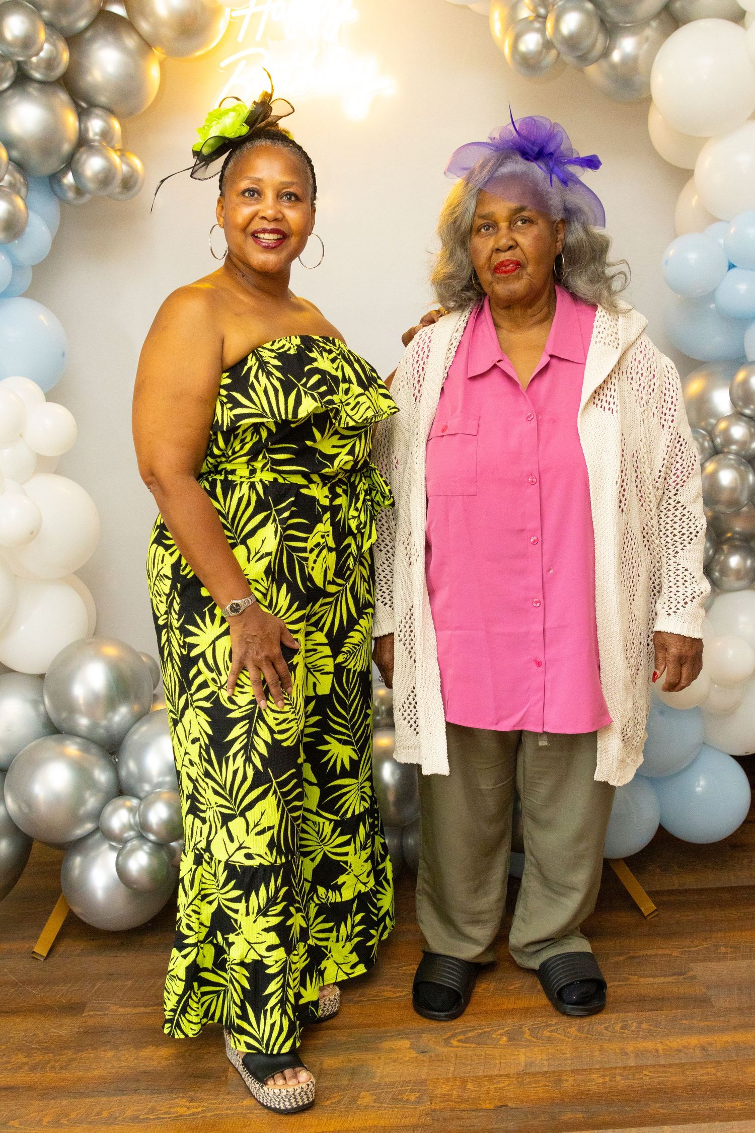 People at a party posing in front of silver and blue balloon decorations with one in a bright yellow floral dress.