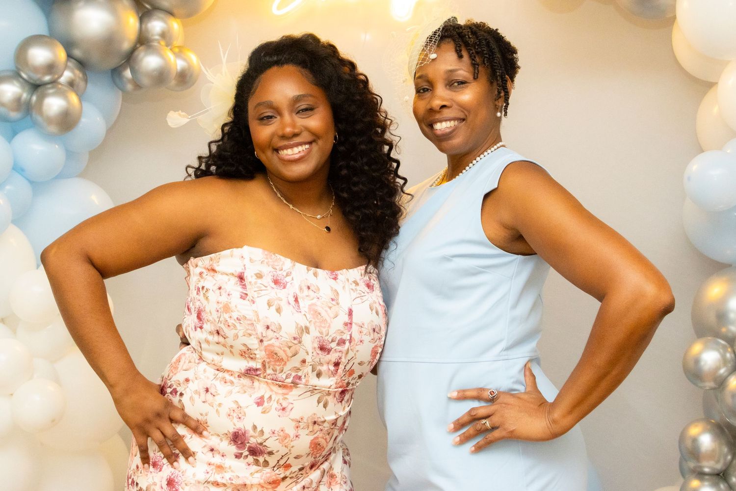 Two smiling friends pose together against a backdrop of silver and blue balloons at a celebration event.