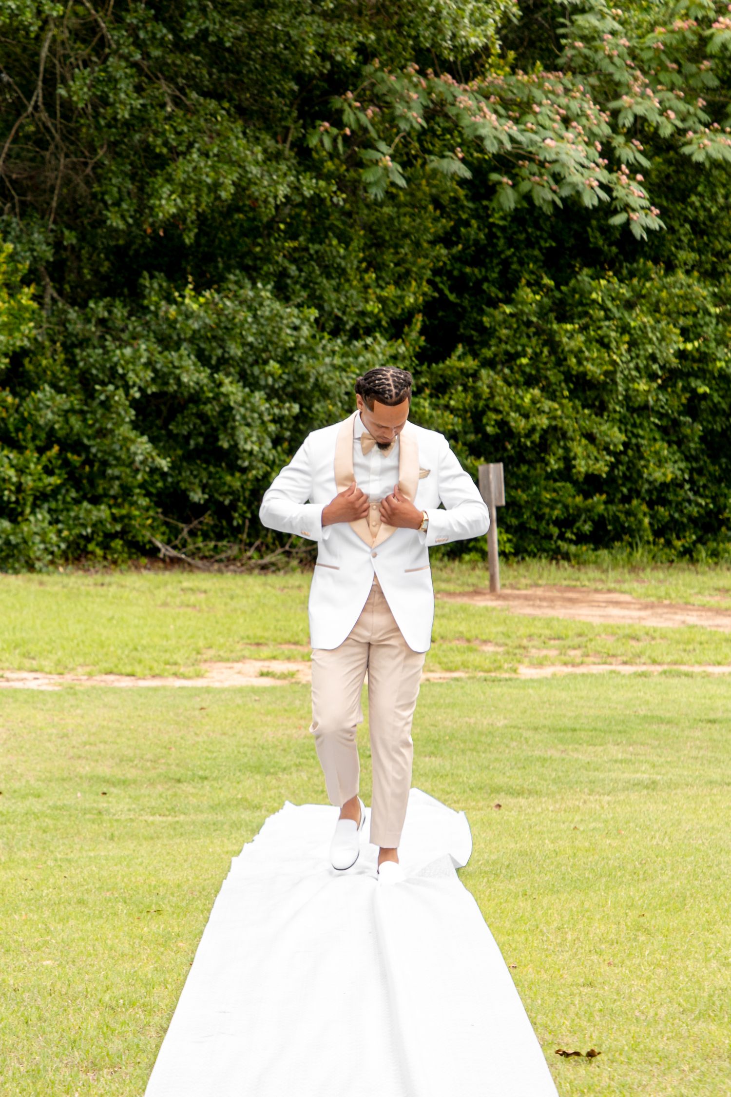 Groom in white blazer and khaki pants walks down white aisle runner toward outdoor wedding ceremony.