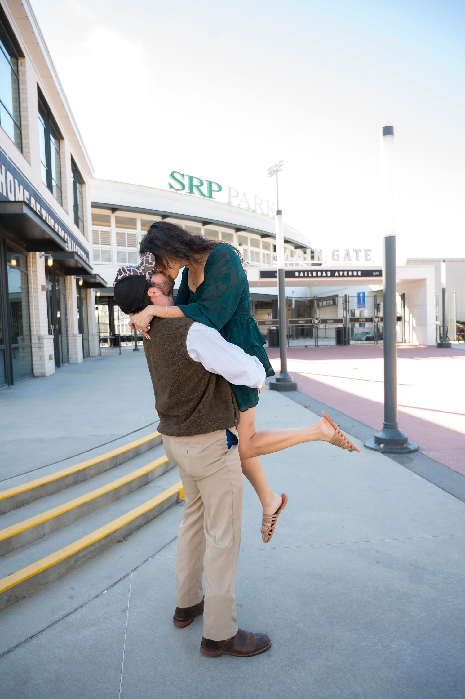 Couple sharing a romantic kiss outside SRP Park baseball stadium, man lifting woman as she kicks up her heel.