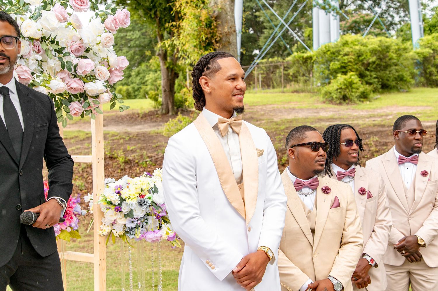 Groom in white tuxedo stands at the altar with groomsmen in tan suits during outdoor wedding ceremony.