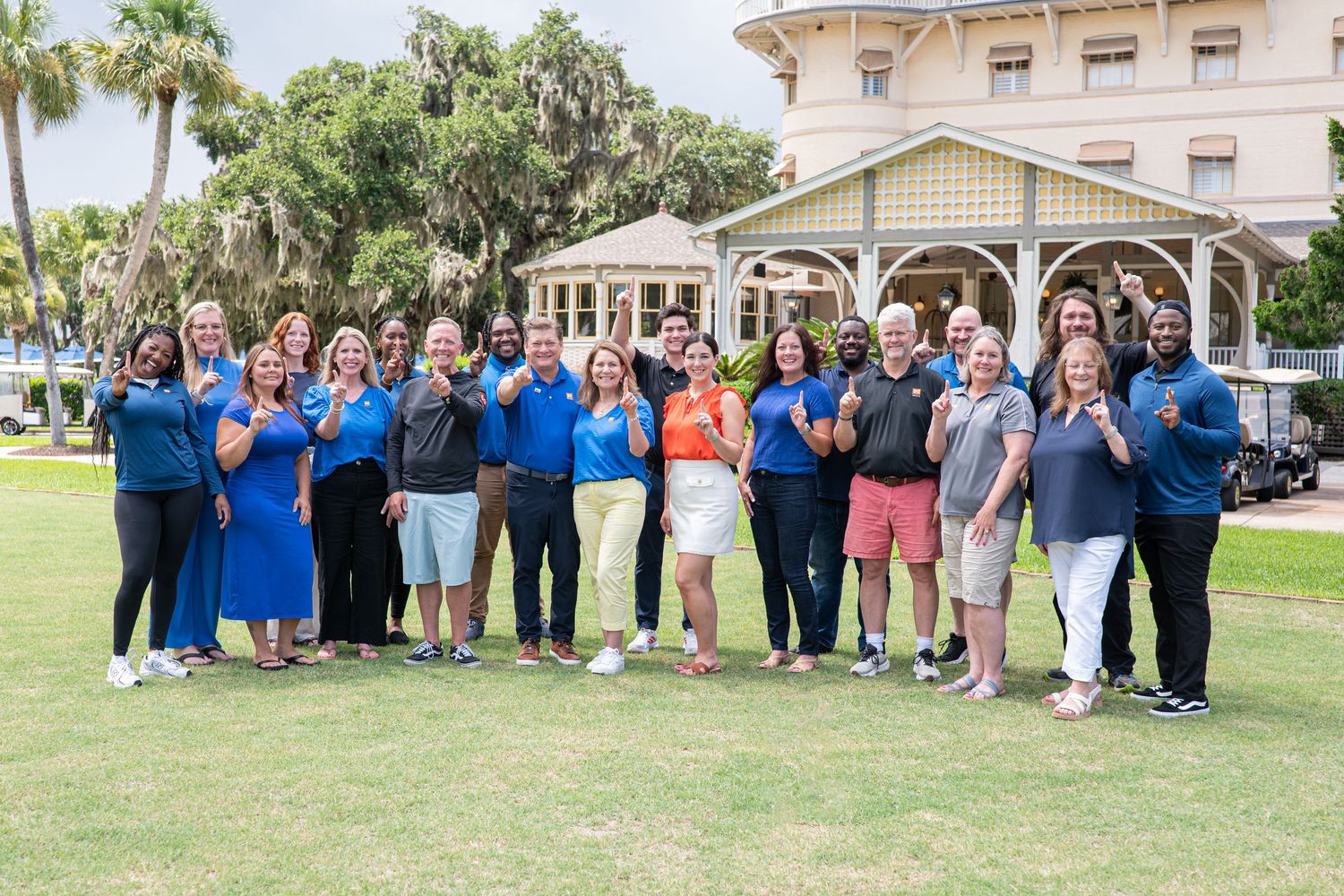 Large group of smiling professionals posing outdoors on golf course lawn near historic yellow building.