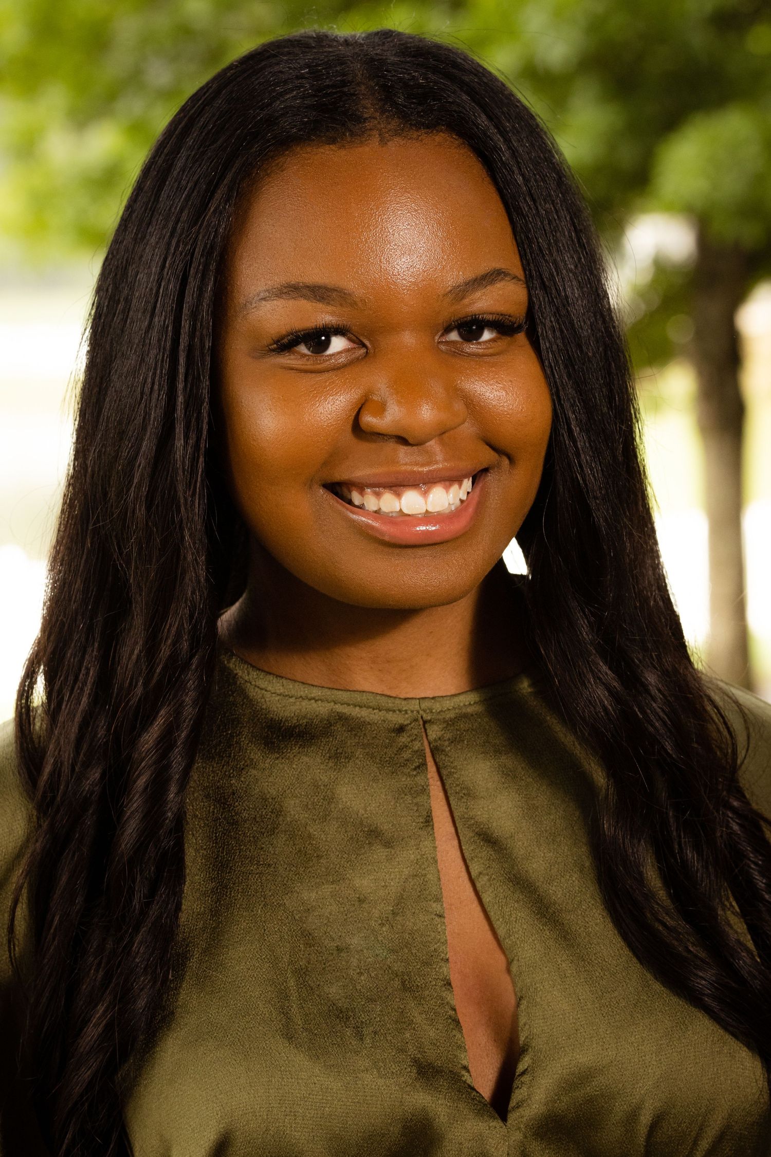 Smiling young Black woman with long hair wearing an olive green top, photographed outdoors with blurred trees.