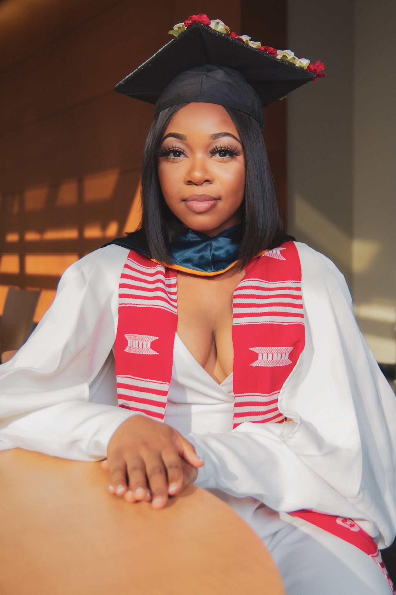 Young woman in white graduation gown, cap with flowers, and red kente stole posing confidently indoors.