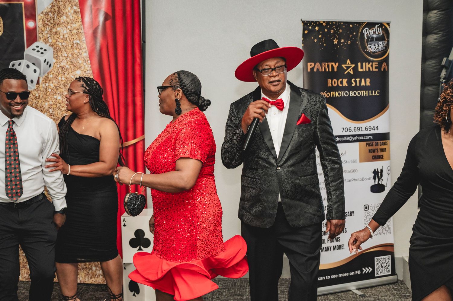Casino themed party with guests in formal black and red attire mingling in front of decorative backdrop.