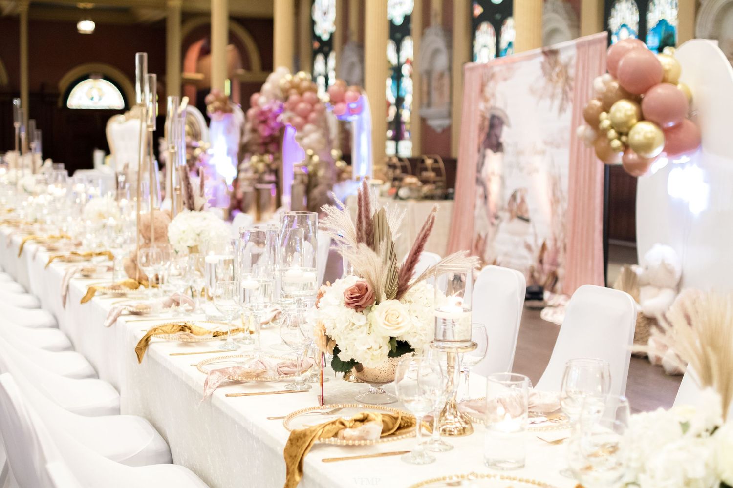 Elegant wedding reception table setting with white linens, floral centerpieces, and golden decor in a luxurious ballroom.