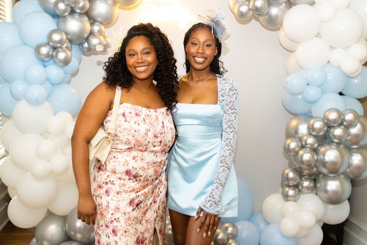 Two friends posing together at a celebration with balloon decorations in blue, white and silver.
