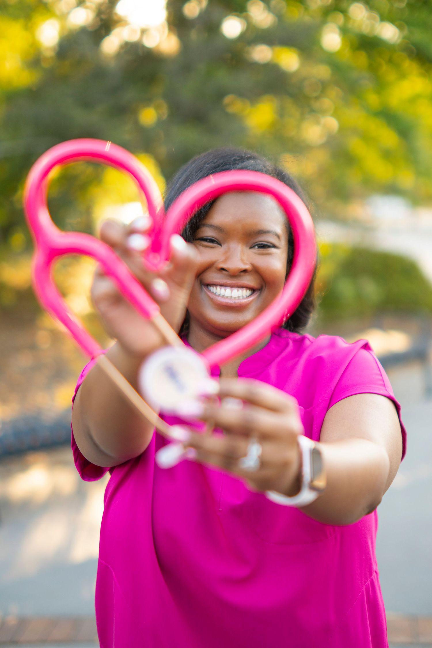 A smiling Black female nurse in pink scrubs holds a pink stethoscope shaped into a heart outdoors.