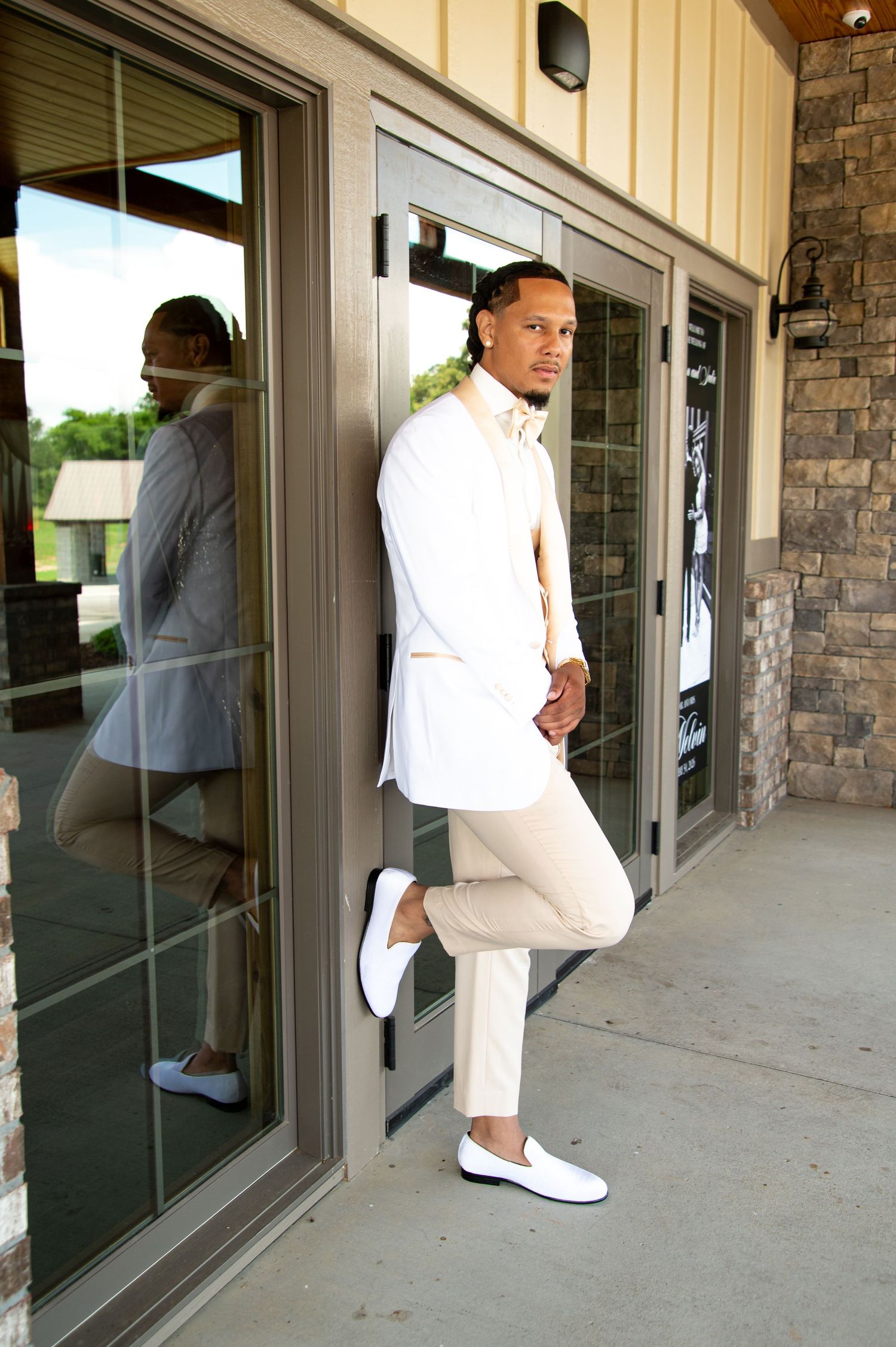 Stylish groom in white shirt and cream pants posing against glass doors at a wedding venue.