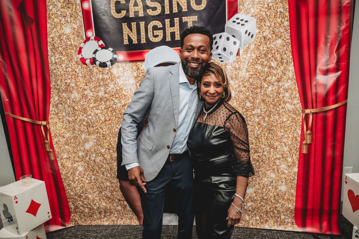 Couple posing in front of a casino-themed photo backdrop with playing cards and dice decorations.