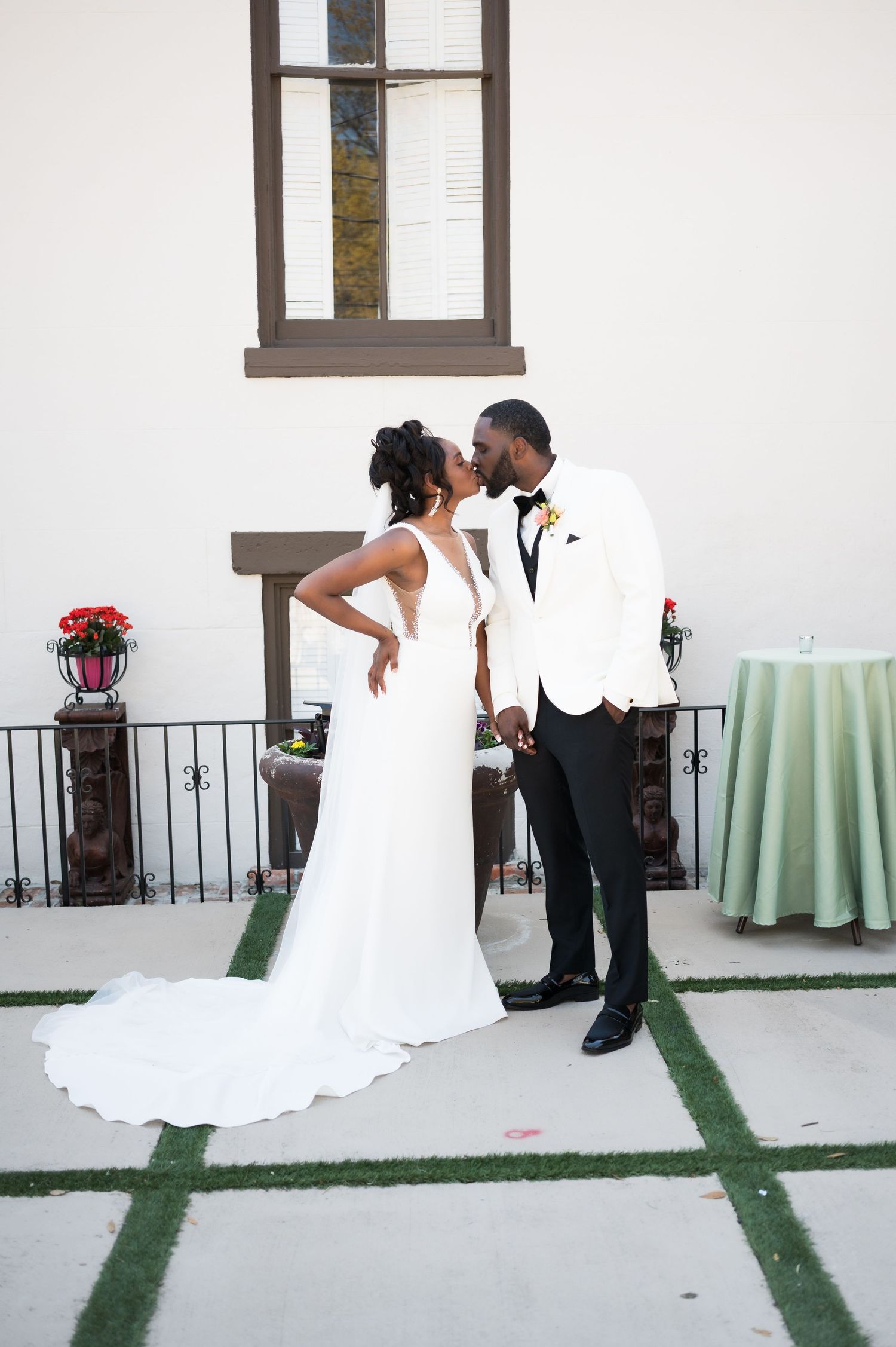 A bride and groom share a kiss outdoors, she in a white gown and he in a white tuxedo jacket with black pants.