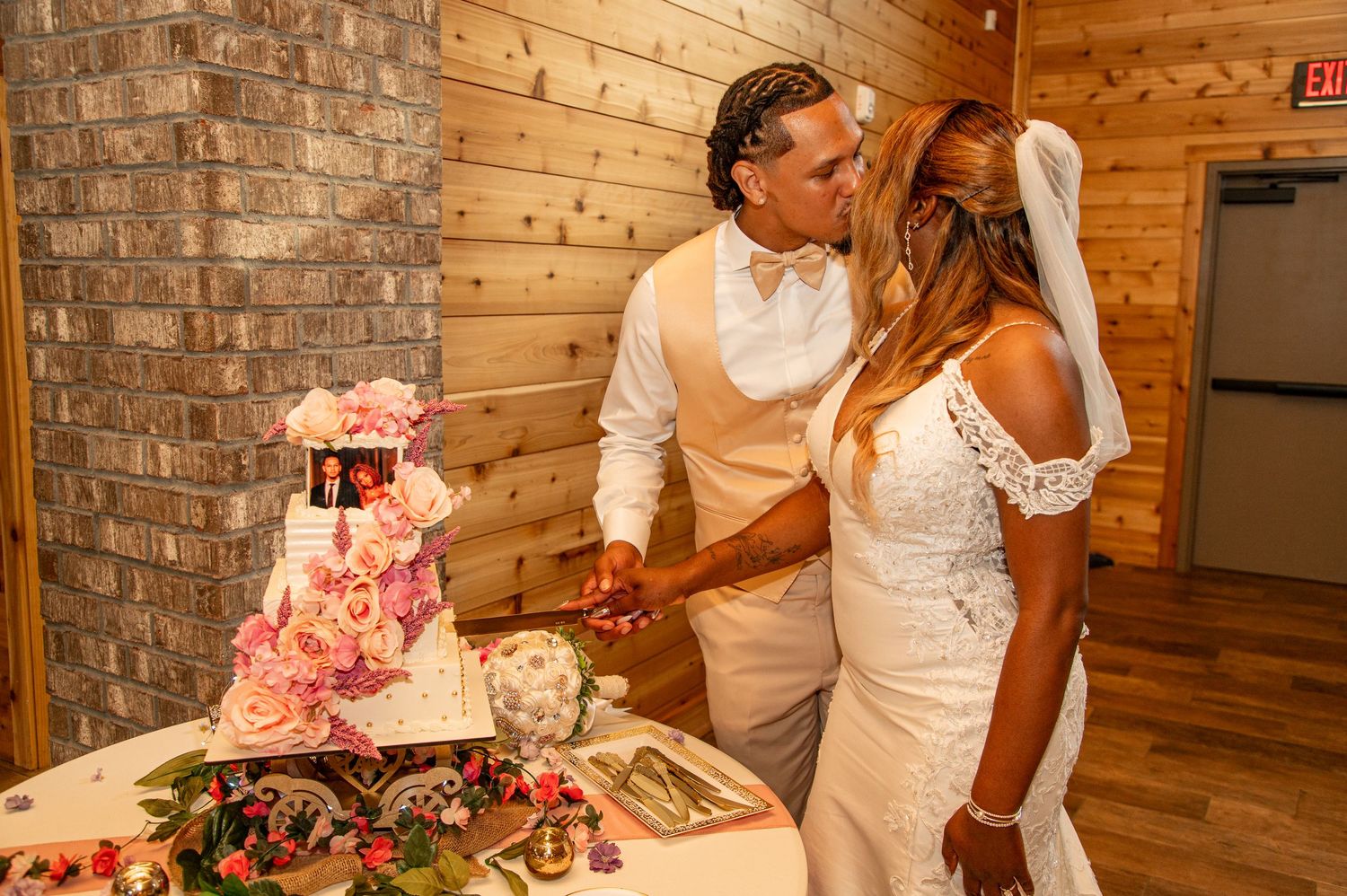 Newlyweds kiss while cutting elegant four-tier floral wedding cake at reception.