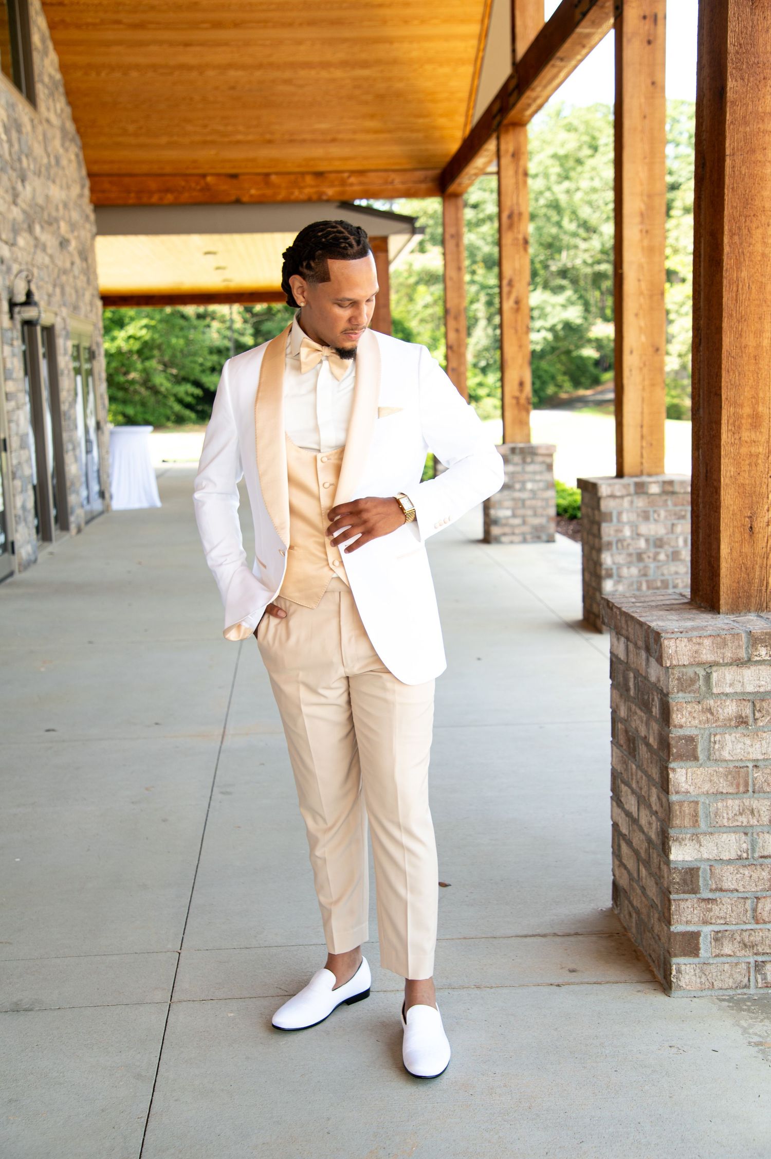 Stylish groom poses in white tuxedo jacket with champagne pants and white loafers outdoors.