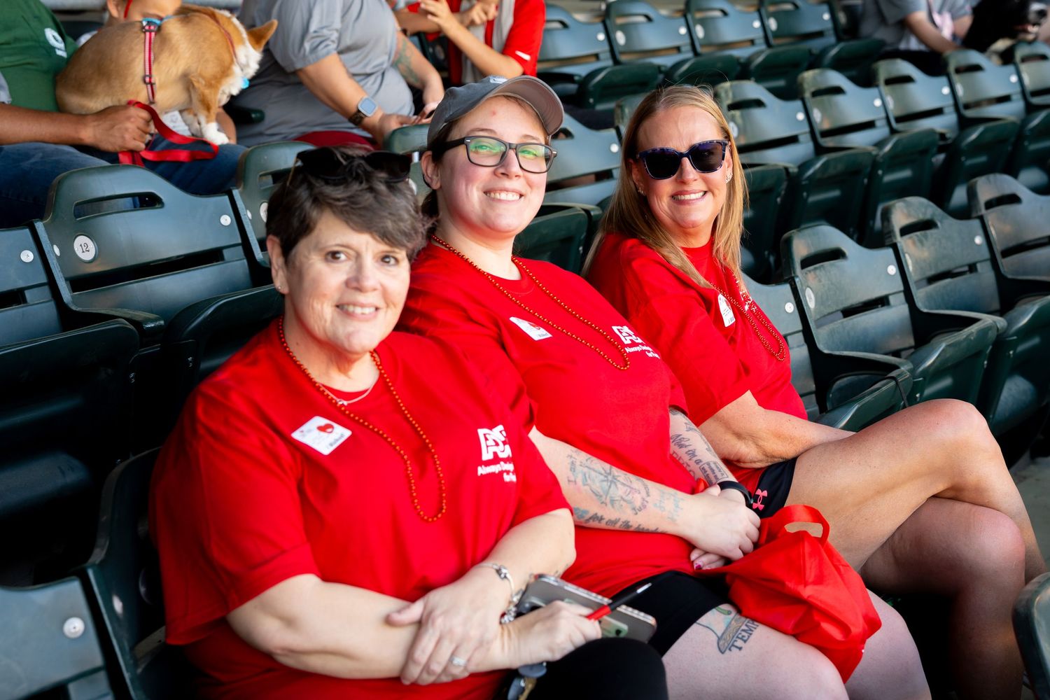 Three staff members in red shirts sit together in stadium seats during an outdoor event.