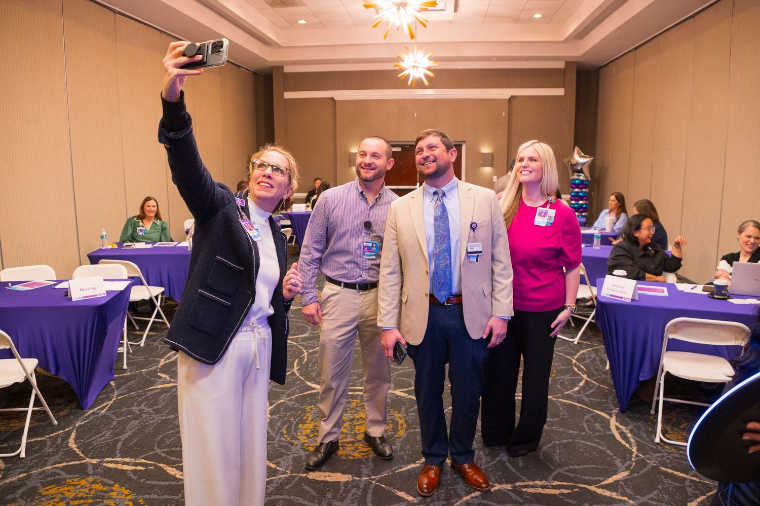 Four professionals pose for a selfie in a conference room with purple-draped tables during a business event.