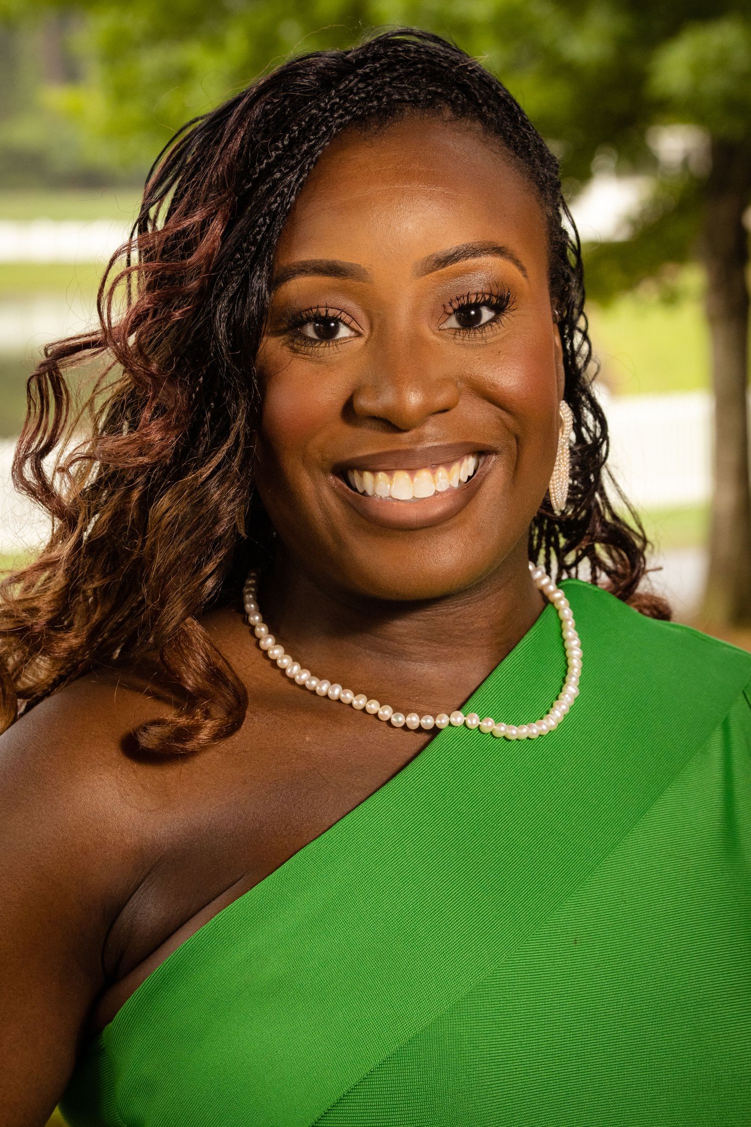 Smiling woman in green one-shoulder dress with pearl necklace poses outdoors for professional portrait photo.