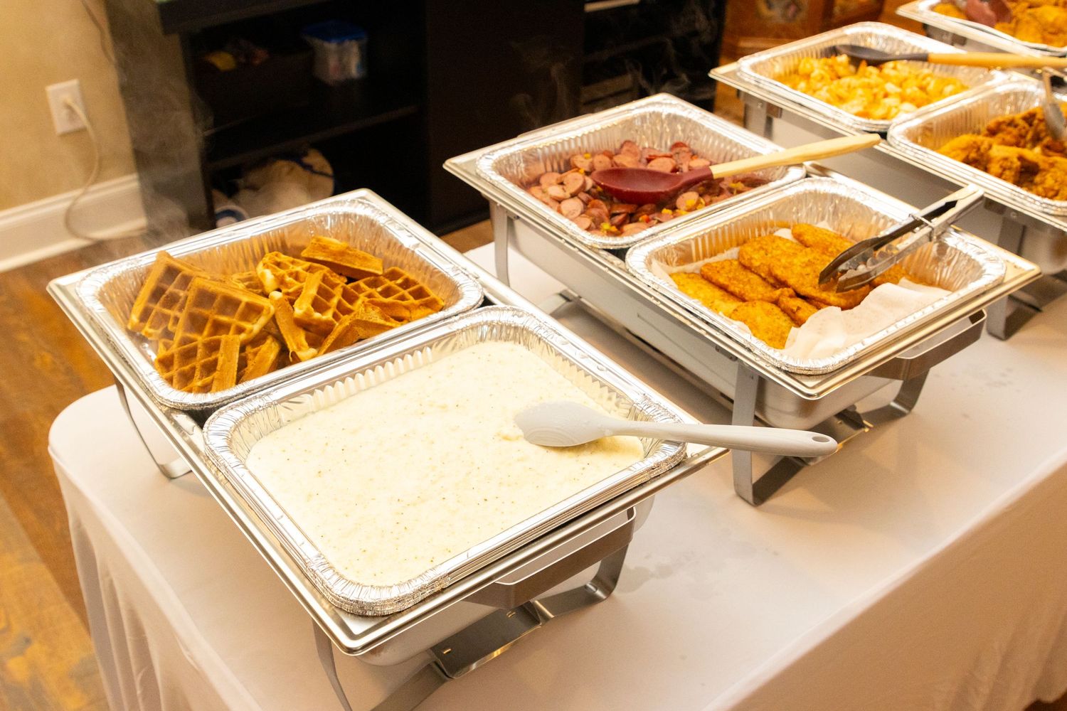 Stainless steel buffet chafing dishes with Asian cuisine including noodles, rice and various entrees on white tablecloth.