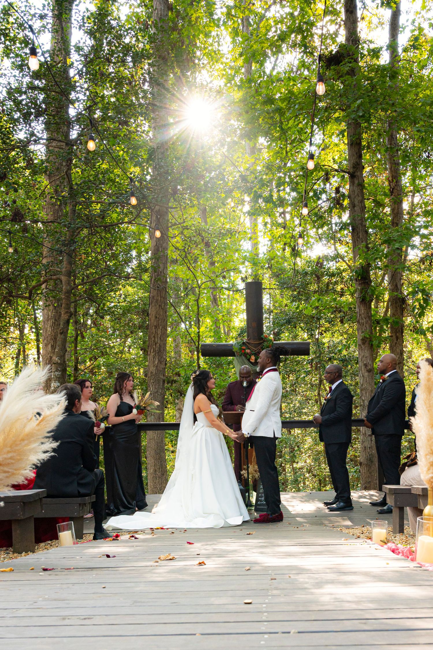 A wedding ceremony takes place on a wooden deck surrounded by sunlit trees in a forest setting.