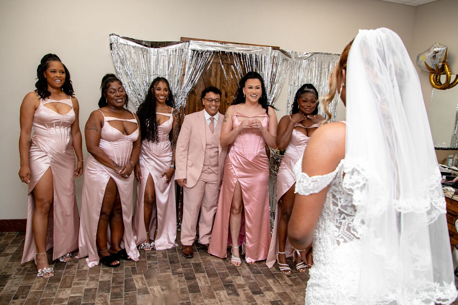 Groom in pink suit poses with bridesmaids in pink satin gowns as bride in white wedding dress looks on.