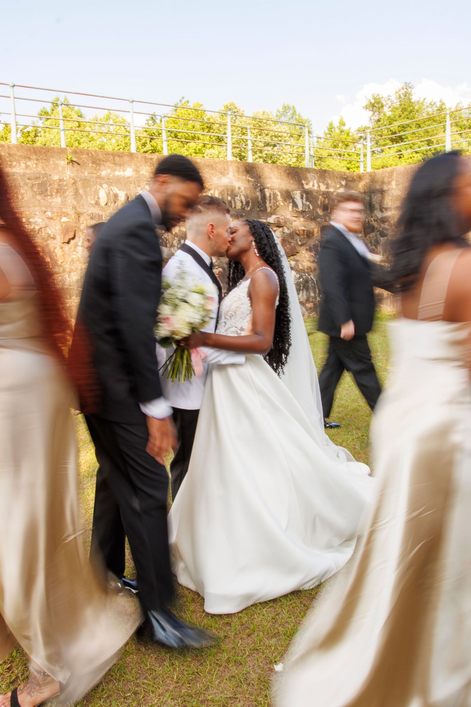 Bride and groom share a kiss surrounded by blurred wedding party in golden gowns and dark suits outdoors.