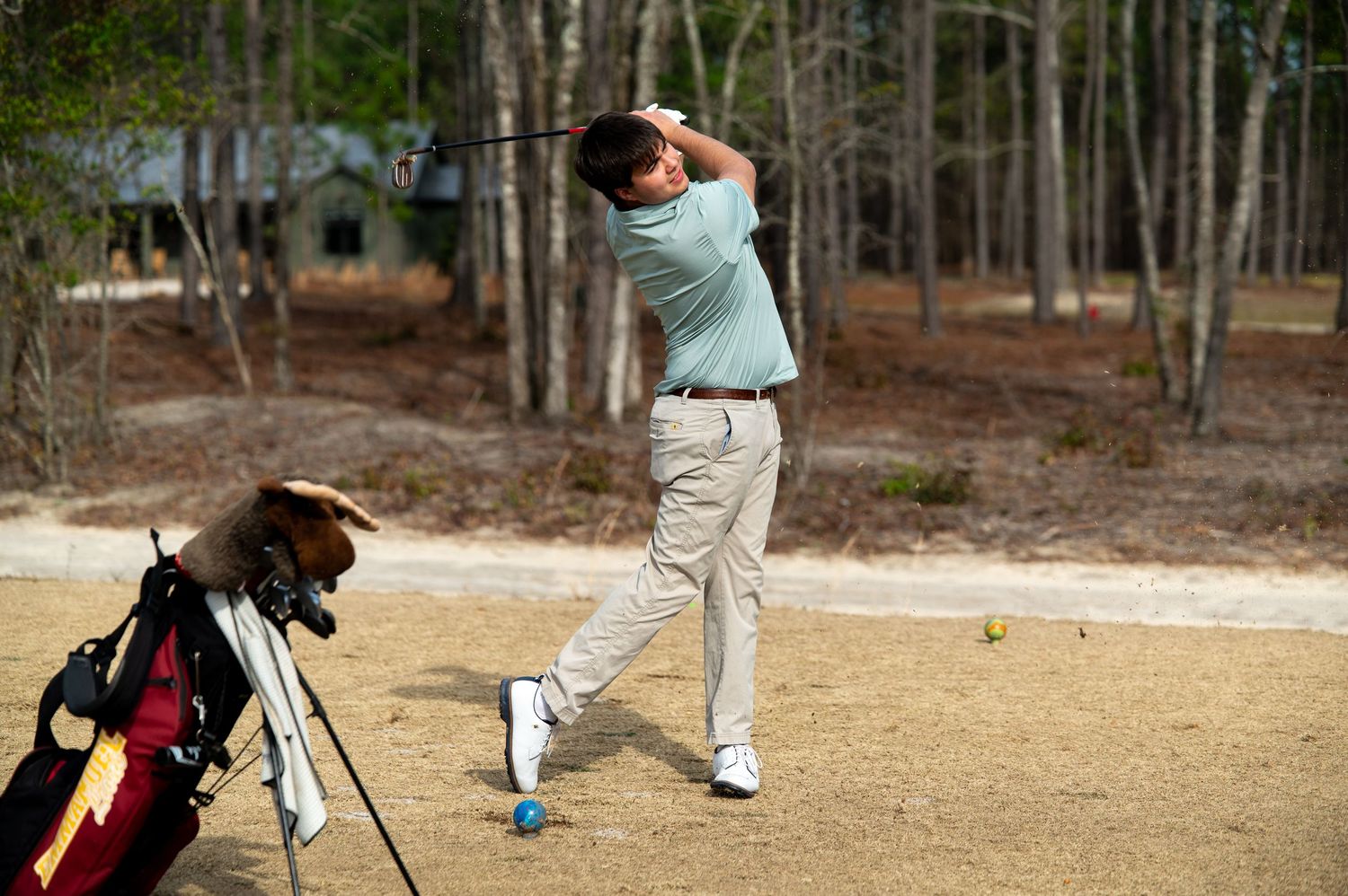 A golfer takes a swing on a sandy golf course surrounded by pine trees in the background.