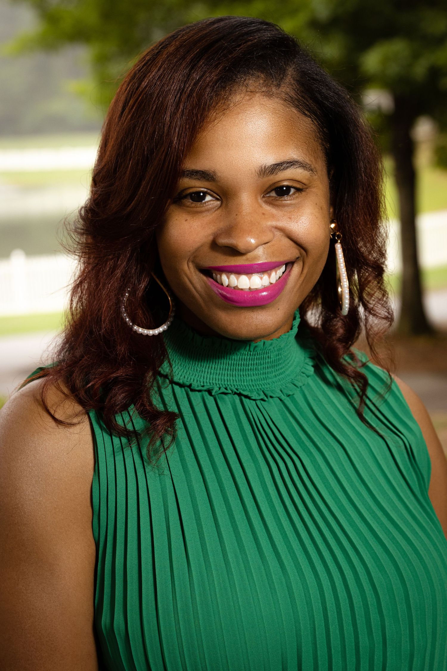 Smiling young Black woman in green turtleneck top with hoop earrings and pink lipstick posing outdoors.