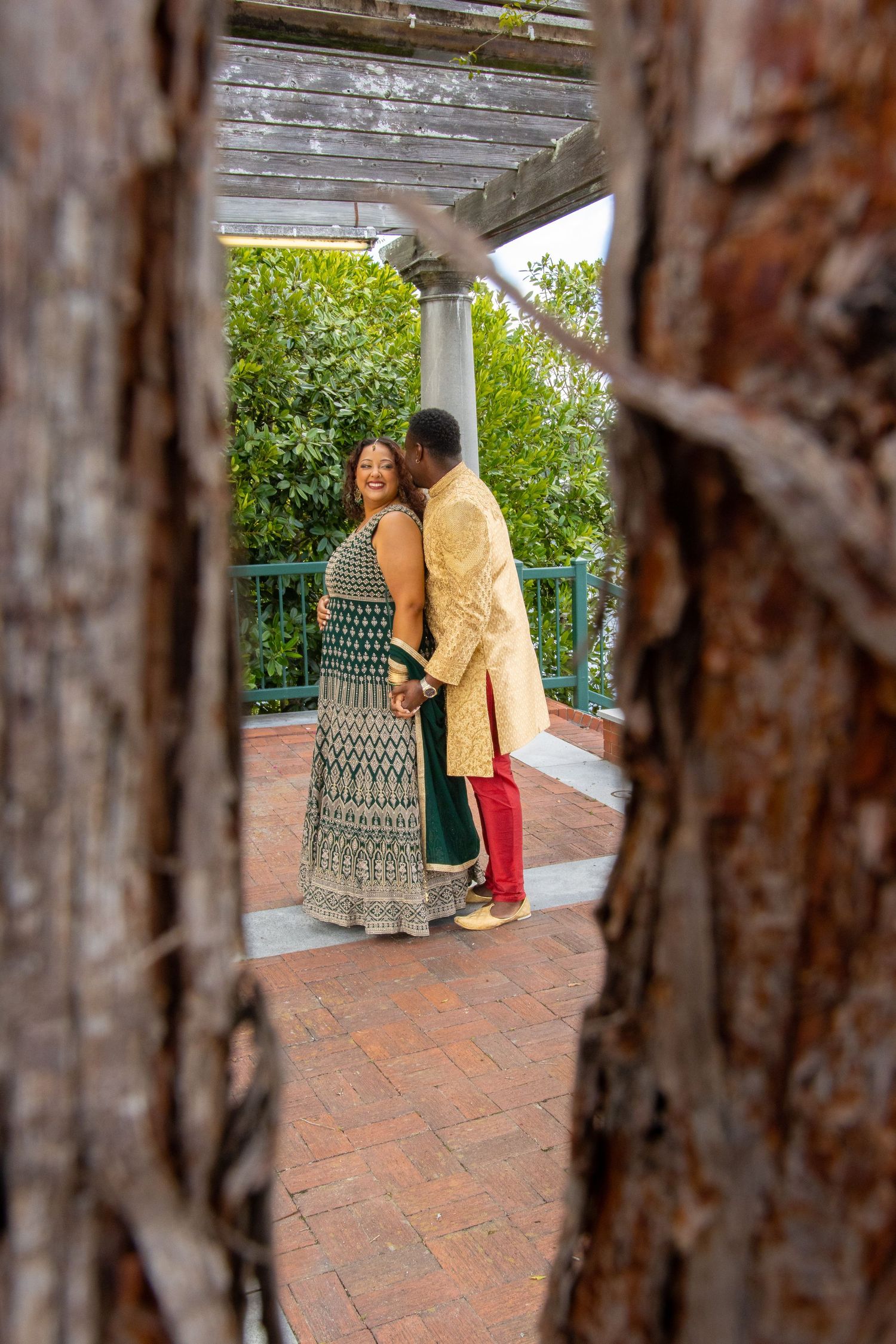 A couple in traditional Indian attire embracing on a terrace with brick flooring and columns framed through tree bark.