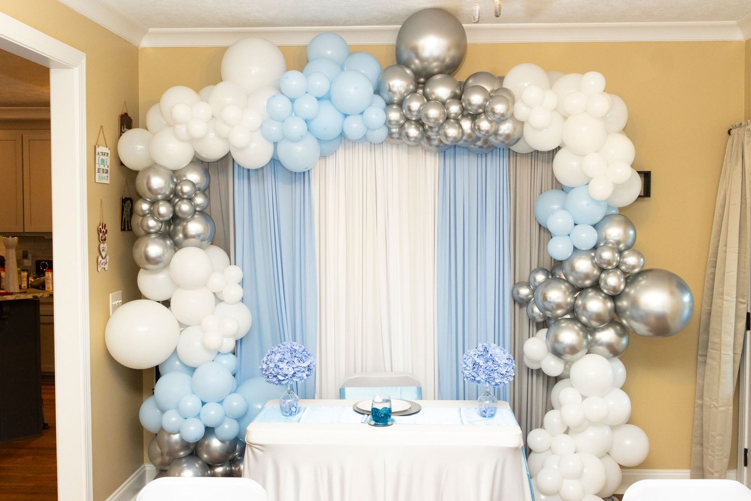 Balloon arch decoration in white, light blue, and silver colors frames a table setting for a party or celebration.