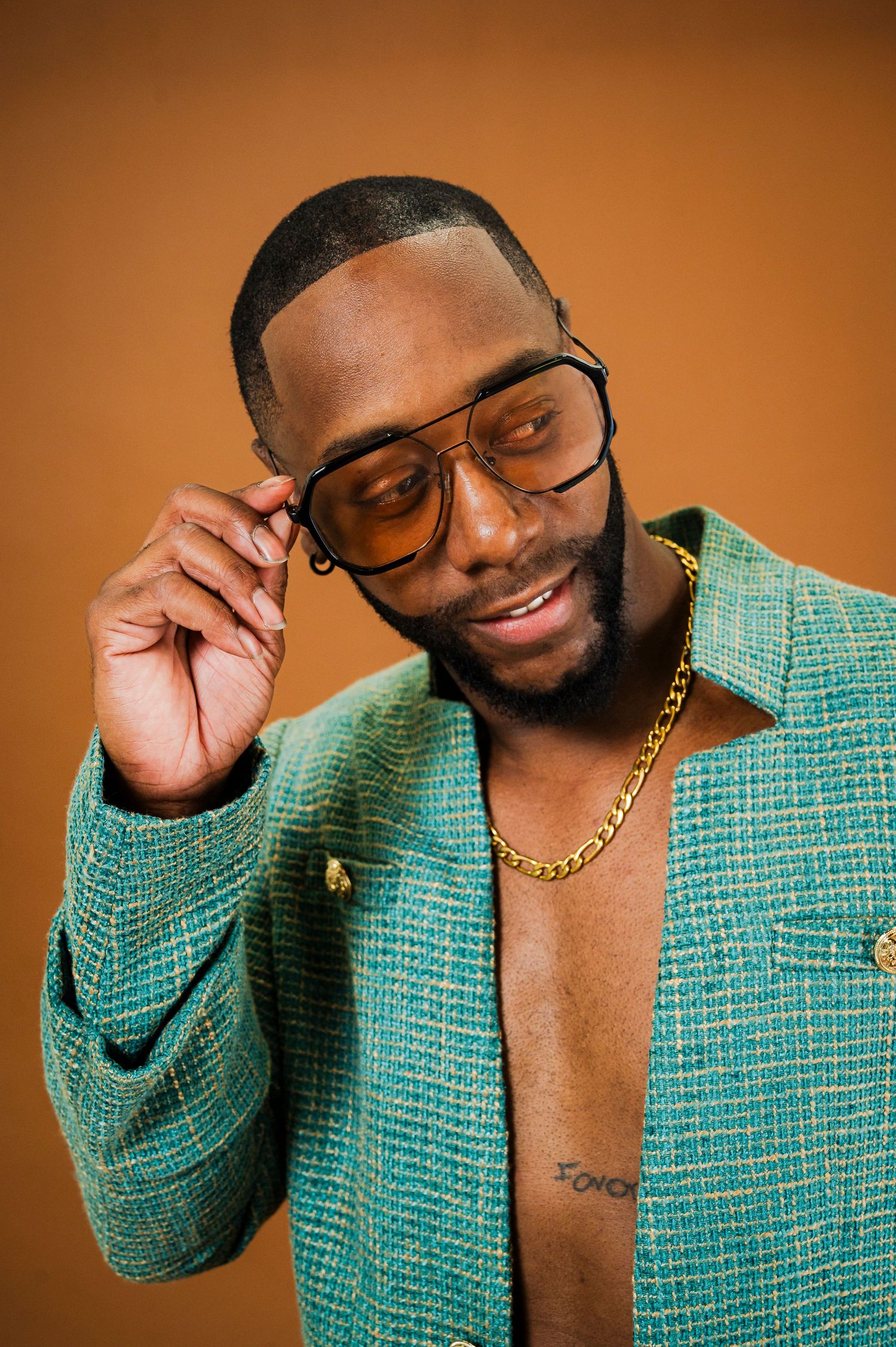 A person wearing a turquoise tweed jacket and gold chain poses playfully in a series of photos against a brown backdrop.