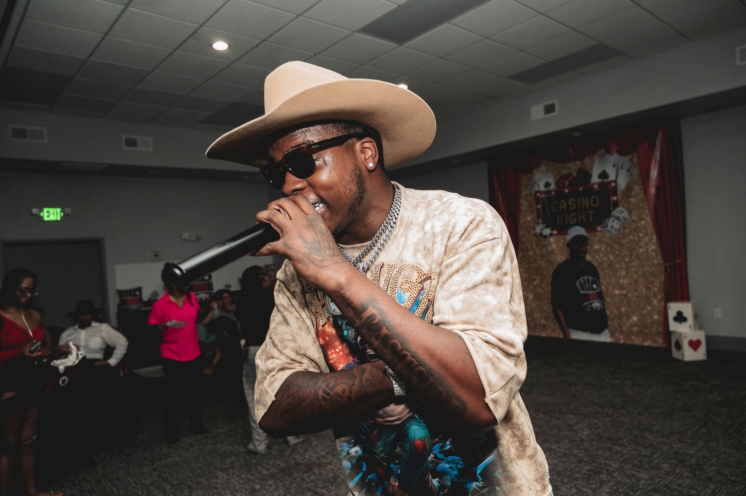 Performer in cowboy hat and sunglasses performs with microphone in indoor venue.