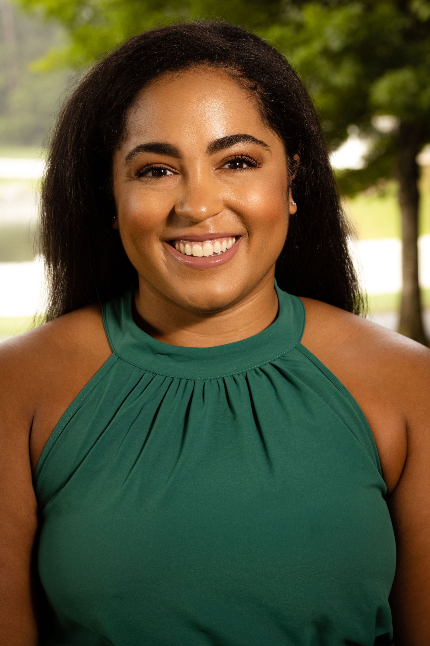 Smiling young woman in a green halter top poses outdoors with lush green trees in the background.