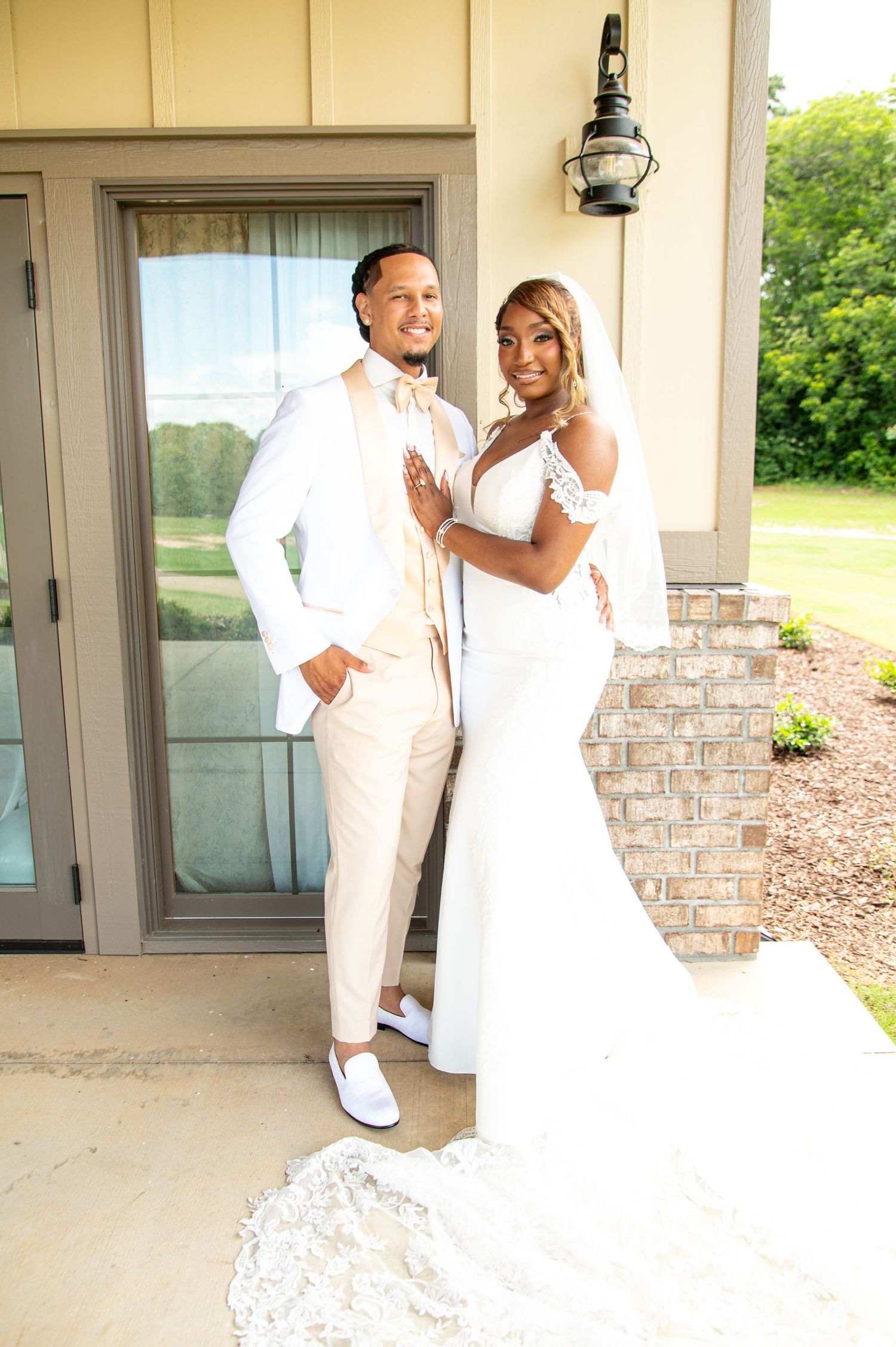 Elegant bride and groom posing together outside venue, wearing white wedding attire with lace details and beige suit.
