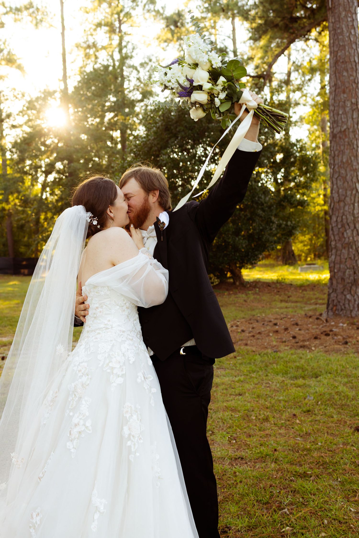 A bride and groom share a kiss outdoors at golden hour, as the groom raises a white floral bouquet in celebration.