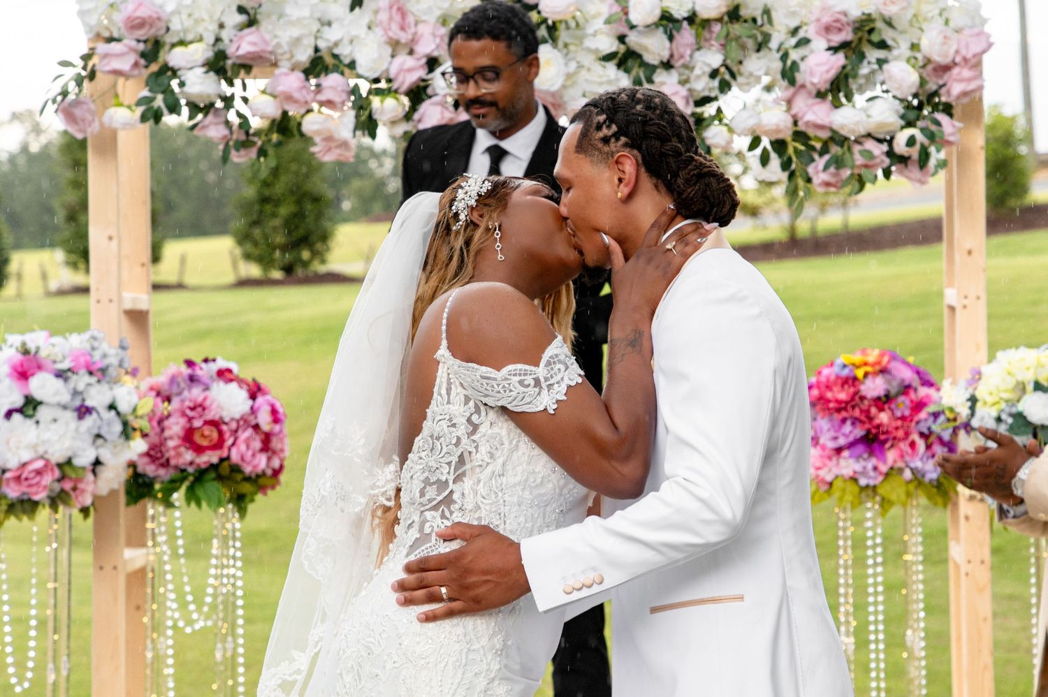 Newlyweds share first kiss under pink floral arch at outdoor wedding ceremony on lush green lawn.