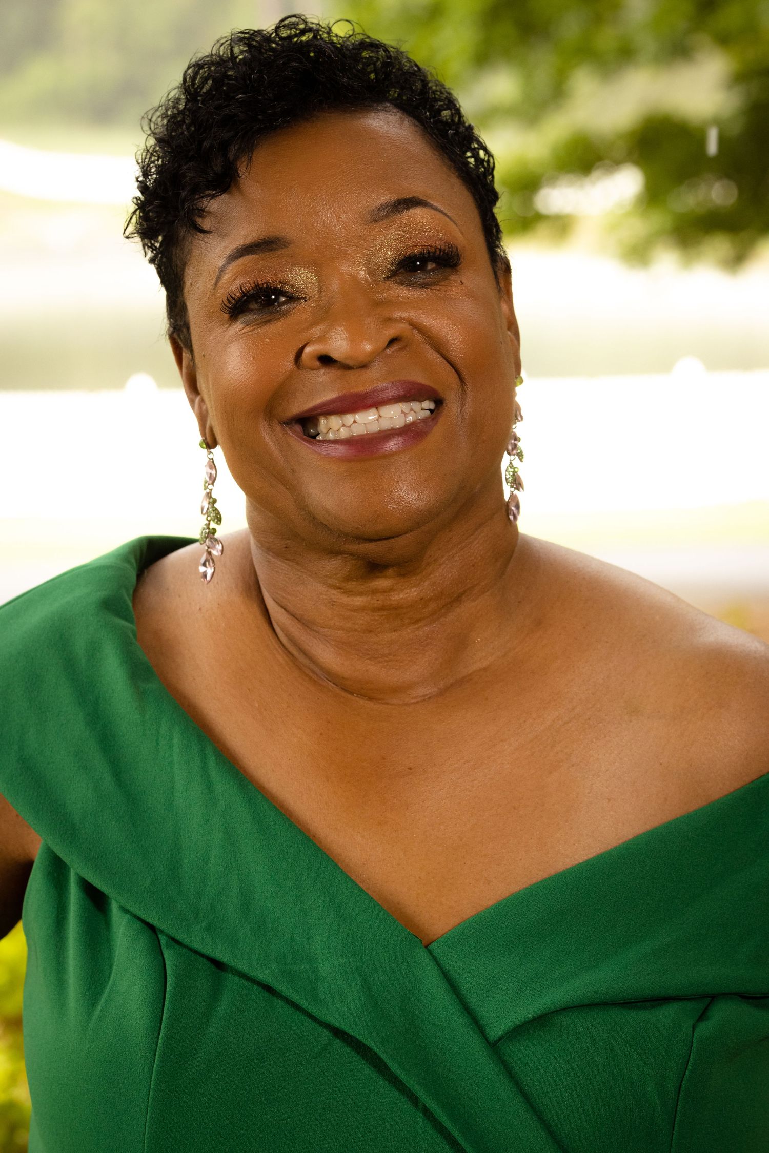 Smiling older Black woman wearing a green off-shoulder dress and drop earrings, posing outdoors with greenery in background.