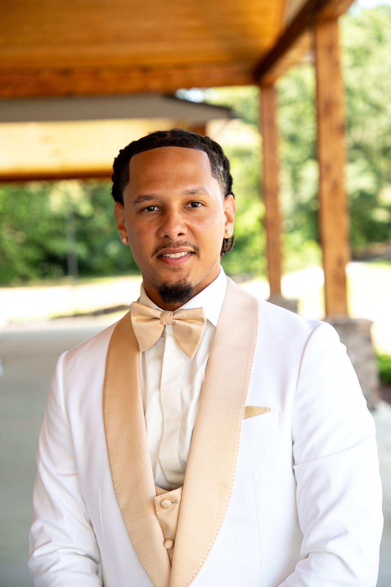 Handsome groom in white tuxedo with champagne bow tie and pocket square smiling at outdoor wedding venue.
