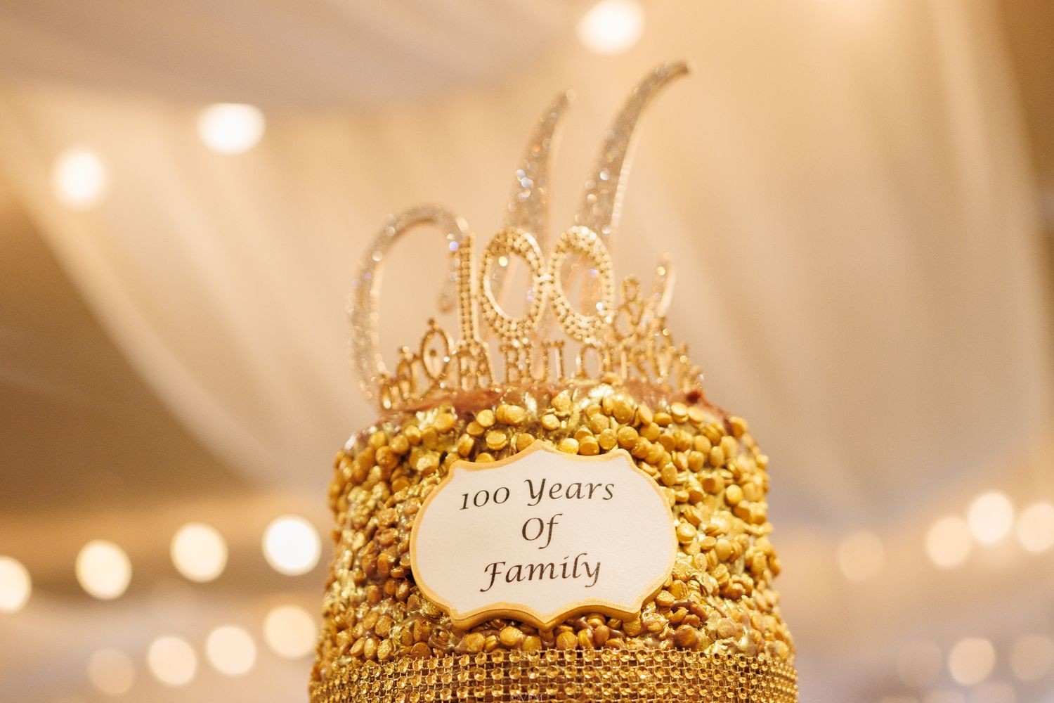 Elegant gold crown cake topper with the phrase 'Joy of family' displayed at a wedding reception with bokeh lights.