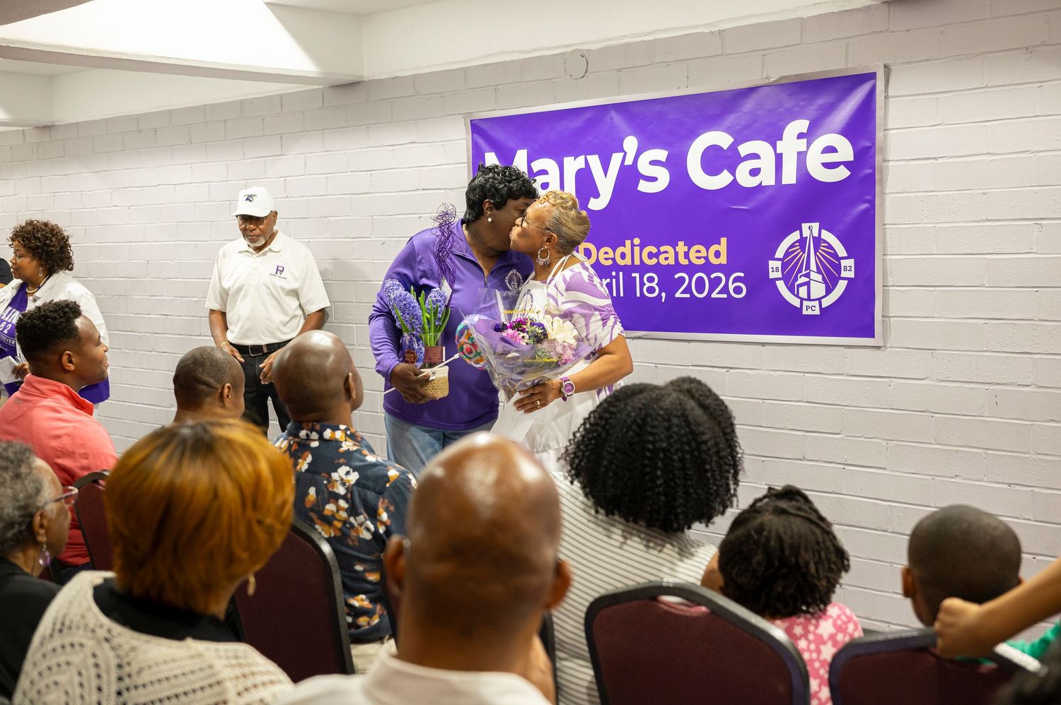 Two people embrace at the dedication of Mary's Cafe, April 18, 2026, surrounded by seated guests in a community hall.